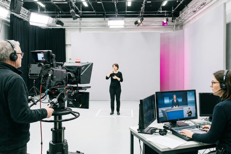 Fernsehstudio mit Kamera, Regisseurin am Mischpult und Gebärdensprachdolmetscherin vor neutraler Wand für barrierefreies TV