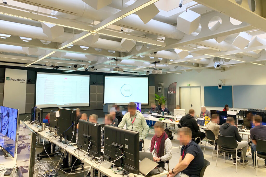 Konferenzraum mit mehreren Arbeitsplätzen, Monitoren und Laptops beim DTVP Plugfest in Berlin, Teilnehmer arbeiten am Setup