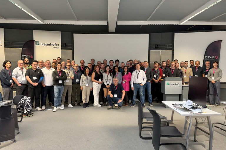 Gruppenfoto von Teilnehmenden beim DTVP Plugfest in Berlin vor Fraunhofer FOKUS Leinwand und Bannern, Konferenzraum, Namensschilder sichtbar