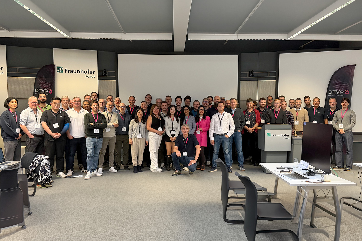 Gruppenfoto von Teilnehmenden beim DTVP Plugfest in Berlin vor Fraunhofer FOKUS Leinwand und Bannern, Konferenzraum, Namensschilder sichtbar