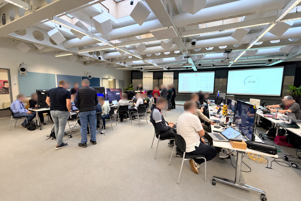 Konferenzraum in Berlin mit Teilnehmern am Hackathon/Plugfest, Laptops, Monitoren und Präsentationsbildschirmen