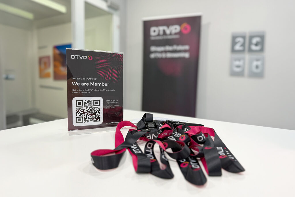Stand mit DTVP-Infotafel, QR-Code und schwarzen/rosa DTVP-Ausweisbändern auf weißer Tischfläche bei Plugfest Berlin