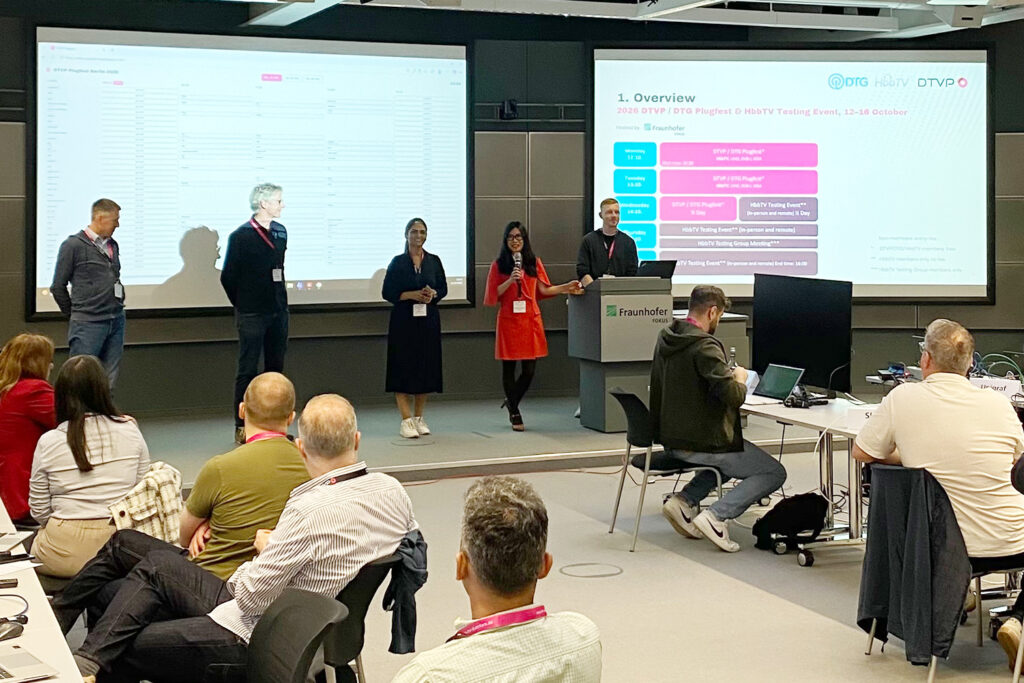 Konferenzraum mit Publikum; fünf Vortragende auf Bühne vor zwei Projektionswänden zeigen Präsentation beim DTVP Plugfest Berlin